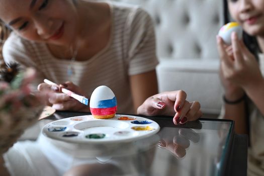 Young woman hands holding paintbrushes and painting on egg while preparing for Easter festival with her daughter.