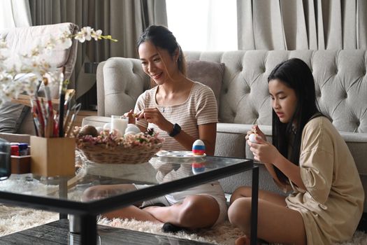 Joyful young woman and mother are sitting in bright living room and coloring Easter eggs together.