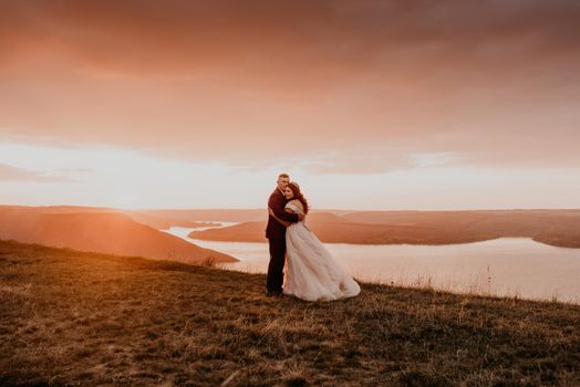 A loving couple wedding newlyweds walk in tsummer in the fall on mountain on cliff above the river. sunset. bride in a white dress with crown on her head. style fashionable women hairstyle makeup