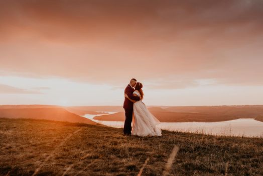 A loving couple wedding newlyweds walk in tsummer in the fall on mountain on cliff above the river. sunset. bride in a white dress with crown on her head. style fashionable women hairstyle makeup