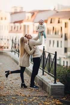 Side view of happy husband and wife with their lovely child. Handsome man holding little baby and beautiful woman hugging him from back. Young family in casual clothes walking together in sunny day.