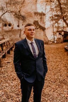 a man in a formal suit the groom stands in the autumn forest he holds his hands in his pockets. rock monastery in bakota