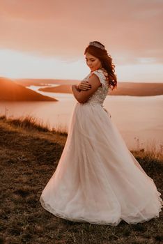young brunette bride in white wedding dress with a crown on her head stands on cliff against the background of the river and islands After sunset.style fashionable women hairstyle makeup