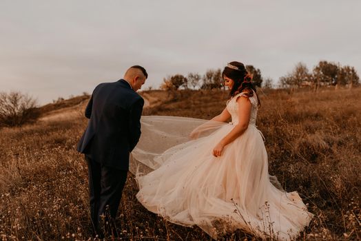 groom cleans mud from bride wedding dress in middle of field in tall grass in autumn. luxury fluffy chiffon dress with motes. spoiled dirty wedding dress