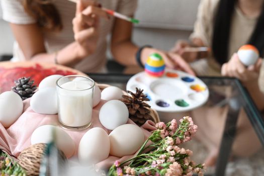 Wicker basket full of eggs and flowers on table in living room with mother and daughter painting Easter egg in background.