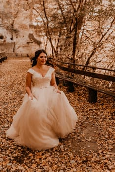 woman bride in white wedding dress with hairstyle makeup and crown on her head walks through autumn forest on fallen orange leaves