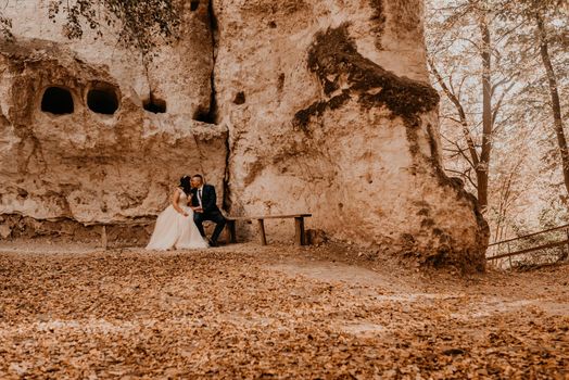 wedding couple in love man and woman sitting on a bench under rock monastery bakota in autumn forest against background of stone rocks. groom suit and bride dress with crown on head