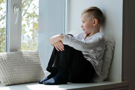 Lonely sad depressed school boy toddler sitting on a windowsill in the house enjoying sunny weather