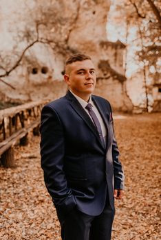 a man in a formal suit the groom stands in the autumn forest he holds his hands in his pockets. rock monastery in bakota