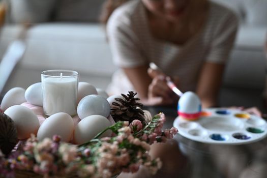 Close up view wicker basket full of eggs, candle and flowers on table with young woman painting Easter egg in background.