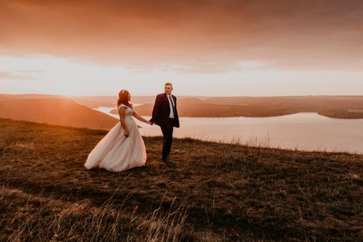 A loving couple wedding newlyweds walk in tsummer in the fall on mountain on cliff above the river. sunset. bride in a white dress with crown on her head. style fashionable women hairstyle makeup