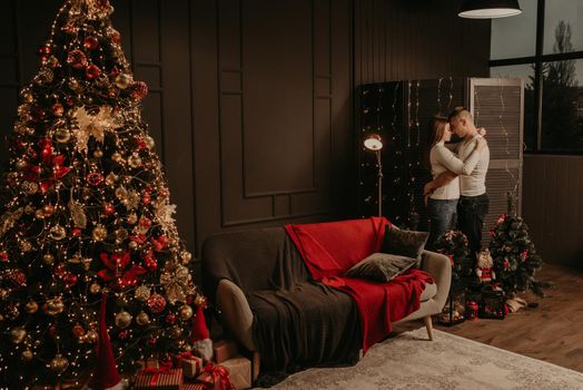 love young couple man and woman hugging kissing near a Christmas tree. decorated house for New Year. Christmas morning. apartment interior. Valentine's Day celebration