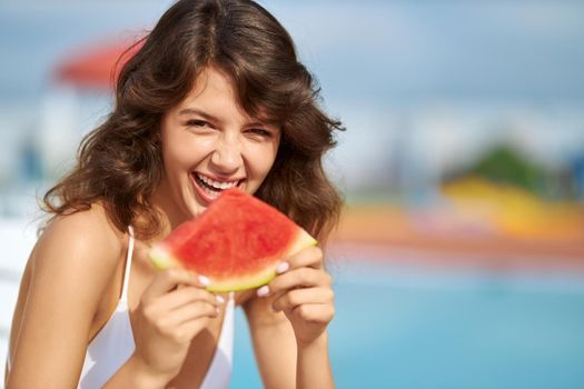Cheerful brunette female eating juicy slice of watermelon outdoors. Portrait view of charming joyful girl holding ripe watermelon segment, looking at camera at summer midday. Concept of satisfaction.