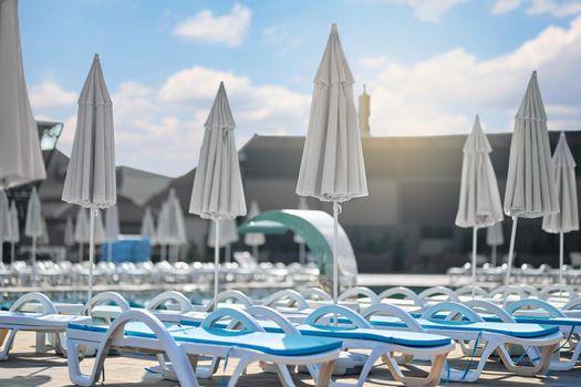 Empty luxury resort with swimming pool, blue loungers and folded umbrellas at sunset. Side view of swimming pool facilities with chaise lounge chairs and blue sky on background. Concept of resting.