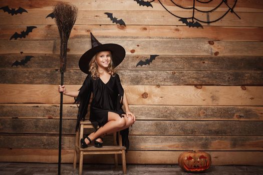Halloween Witch concept - Full-length shot of little caucasian witch child posing with magic broomstick over bat and spider web background