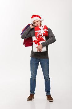 Christmas Concept - young handsome man with beard holding heavy presents and shopping bags with exhausted facial expression on white background.