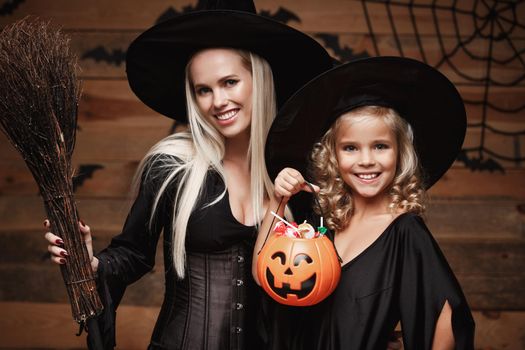 Halloween Concept: beautiful caucasian mother and her daughter in witch costumes celebrating Halloween with sharing Halloween candy and sweet over bats and spider web on Wooden studio background.