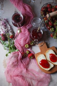 Summer breakfast concept with srawberry jam with bread and fresh harvested berries in basket with pink flowers and cloth, top view, flat lay, selective focus.