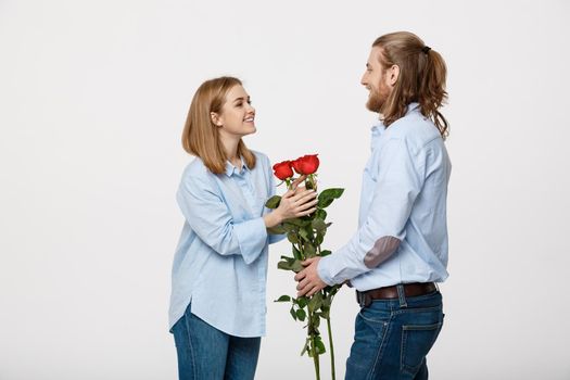 Portrait of handsome elegant guy is surprising his beautiful girlfriend with red roses and smiling over white isolated background .
