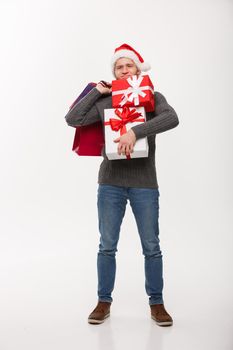 Christmas Concept - young handsome man with beard holding heavy presents and shopping bags with exhausted facial expression on white background.