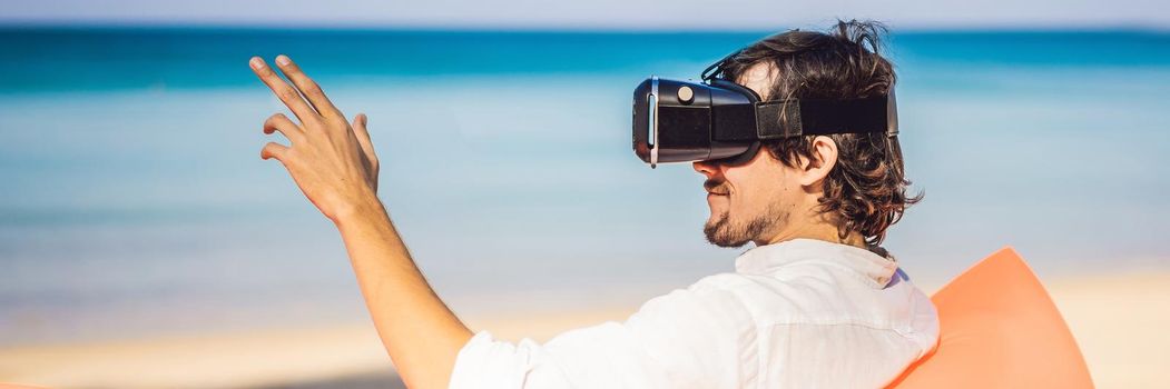 BANNER, LONG FORMAT Summer lifestyle portrait of man sitting on the orange inflatable sofa and uses virtual reality headset on the beach of tropical island. Relaxing and enjoying life on air bed.