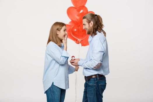 Portrait of Young handsome guy gives a ring to a girl on a white background with red heart air balloons .