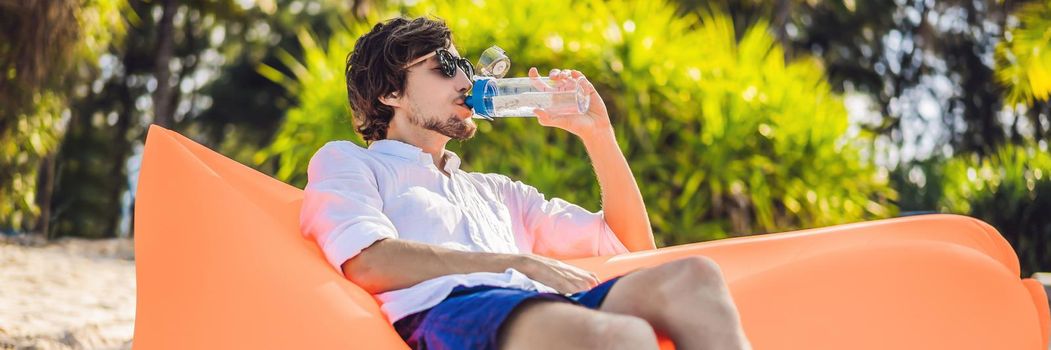 BANNER, LONG FORMAT Summer lifestyle portrait of men sitting on the orange inflatable sofa drinking water on the beach of tropical island. Relaxing and enjoying life on air bed.