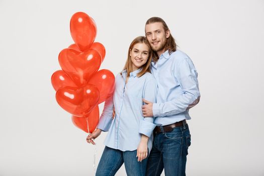 Portrait of young fashionable caucasian couple with balloons heart hugging at each other over isolated white background .