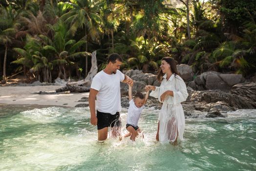 Happy smile european family have rest and run on white sand beach. Father, long black chestnut hair mother, blond baby boy. white cotton clothes. boho dress. t-shorts.Thailand. Aquamarine crystal sea