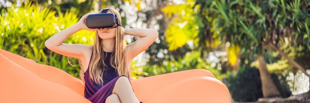 BANNER, LONG FORMAT Summer lifestyle portrait of pretty girl sitting on the orange inflatable sofa and uses virtual reality headset on the beach of tropical island. Relaxing and enjoying life on air bed.