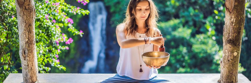 Woman playing on Tibetan singing bowl while sitting on yoga mat against a waterfall. Vintage tonned. Beautiful girl with mala beads meditating. BANNER, LONG FORMAT