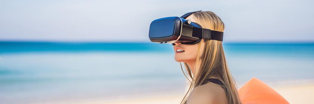 BANNER, LONG FORMAT Summer lifestyle portrait of pretty girl sitting on the orange inflatable sofa and uses virtual reality headset on the beach of tropical island. Relaxing and enjoying life on air bed.