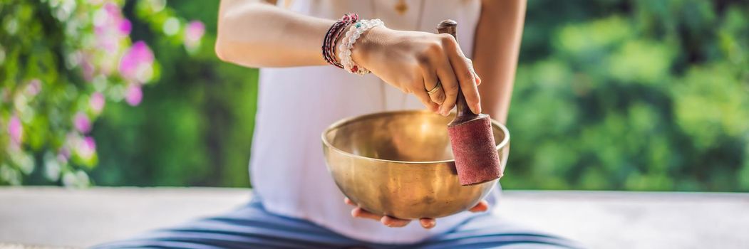 Woman playing on Tibetan singing bowl while sitting on yoga mat against a waterfall. Vintage tonned. Beautiful girl with mala beads meditating. BANNER, LONG FORMAT