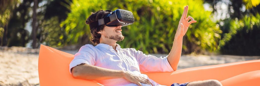 BANNER, LONG FORMAT Summer lifestyle portrait of man sitting on the orange inflatable sofa and uses virtual reality headset on the beach of tropical island. Relaxing and enjoying life on air bed.