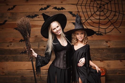 Halloween Concept - cheerful mother and her daughter in witch costumes celebrating Halloween posing with curved pumpkins over bats and spider web on Wooden studio background