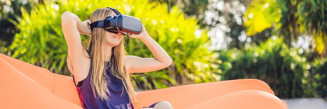 BANNER, LONG FORMAT Summer lifestyle portrait of pretty girl sitting on the orange inflatable sofa and uses virtual reality headset on the beach of tropical island. Relaxing and enjoying life on air bed.