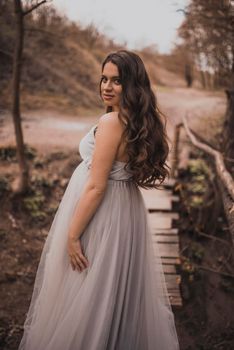 a pregnant black-haired woman with a big belly in a light long dress against a background of mountains walks along a wooden bridge