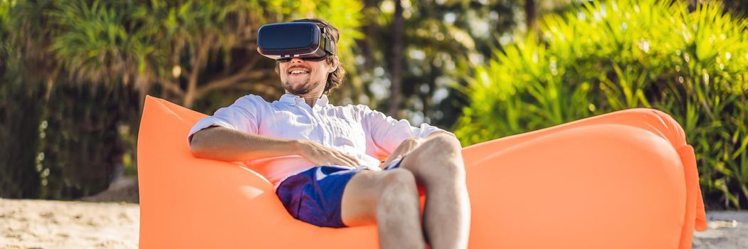 BANNER, LONG FORMAT Summer lifestyle portrait of man sitting on the orange inflatable sofa and uses virtual reality headset on the beach of tropical island. Relaxing and enjoying life on air bed.