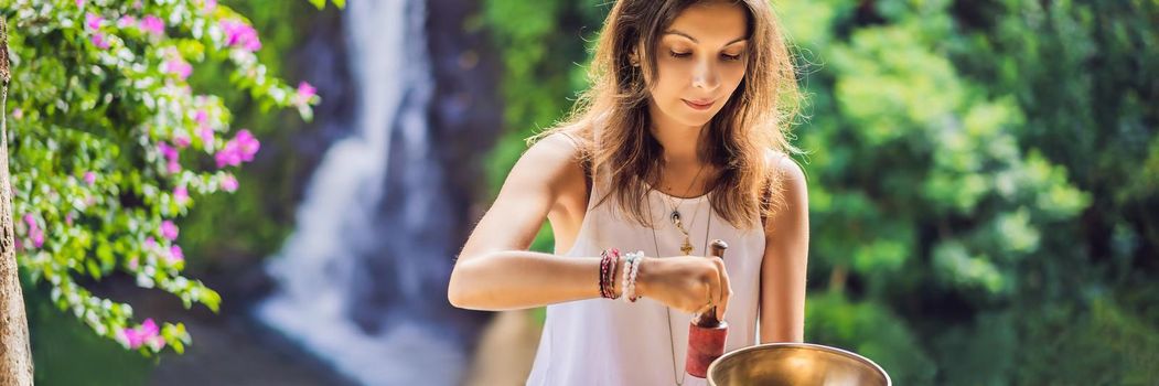 Woman playing on Tibetan singing bowl while sitting on yoga mat against a waterfall. Vintage tonned. Beautiful girl with mala beads meditating. BANNER, LONG FORMAT