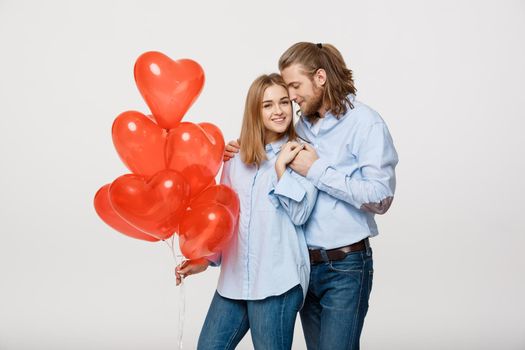 Portrait of young fashionable caucasian couple with balloons heart hugging at each other over isolated white background .