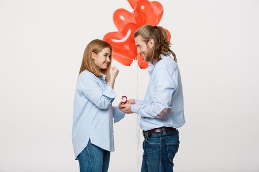 Portrait of Young handsome guy gives a ring to a girl on a white background with red heart air balloons .