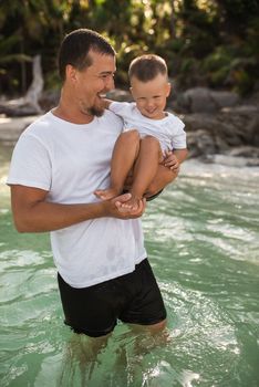 A black-haired young bearded father holds in arms his little child with a blond son.The kid laughs in father arms. family dressed in simple white comfortable clothes.Thailand. Aquamarine crystal sea