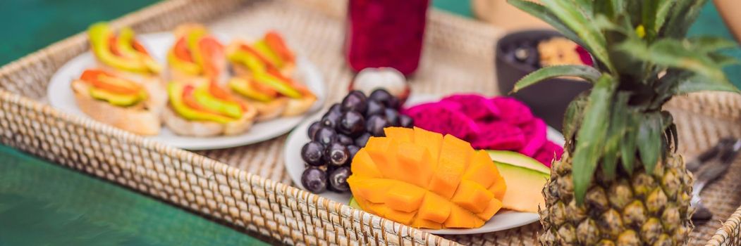 BANNER, LONG FORMAT Breakfast tray in swimming pool, floating breakfast in luxury hotel. Girl relaxing in the pool drinking smoothies and eating fruit plate, smoothie bowl by the hotel pool. Exotic summer diet. Tropical beach lifestyle. Bali Trend.