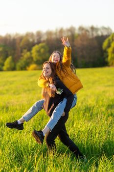 Playful mother giving daughter piggy back ride at green field. Both laughing and look happy. Spring in forest background. Closeup.