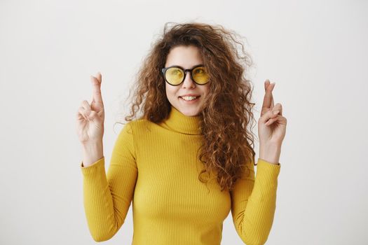 Excited woman in yellow sweater keeping fingers crossed, mouth wide open, waiting for special moment isolated on grey background