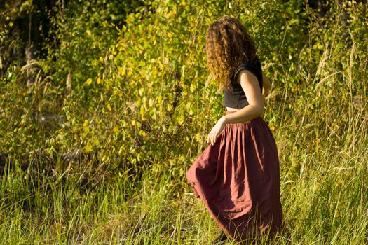 The girl walks across the field. A girl in a bk walks among the plants. Curly girl's autumn walk. The girl is looking for her mood in the forest.