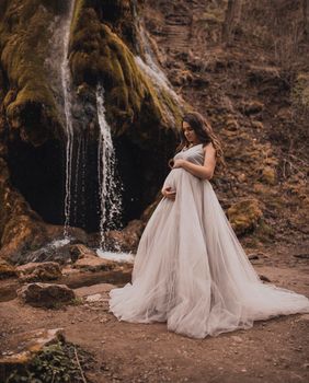 pregnant woman with a big belly in a white long dress in nature on the background of a waterfall
