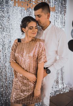 inscription: happy birthday. Young couple lovers man and woman in festive fashionable clothes are hugging. Light casual hairstyle for women with perm.Shiny festive silver photo zone