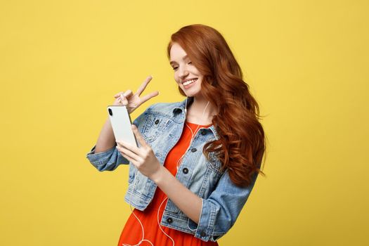 Lifestyle Concept - Portrait ginger hair woman in casual dress using mobile phone for interaction video calling by showing two fingers. Isolated on bright yellow background.