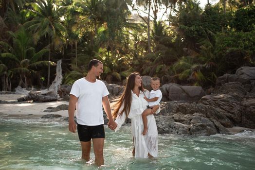 Happy smile european family have rest and run on white sand beach. Father, long black chestnut hair mother, blond baby boy. white cotton clothes. boho dress. t-shorts.Thailand. Aquamarine crystal sea